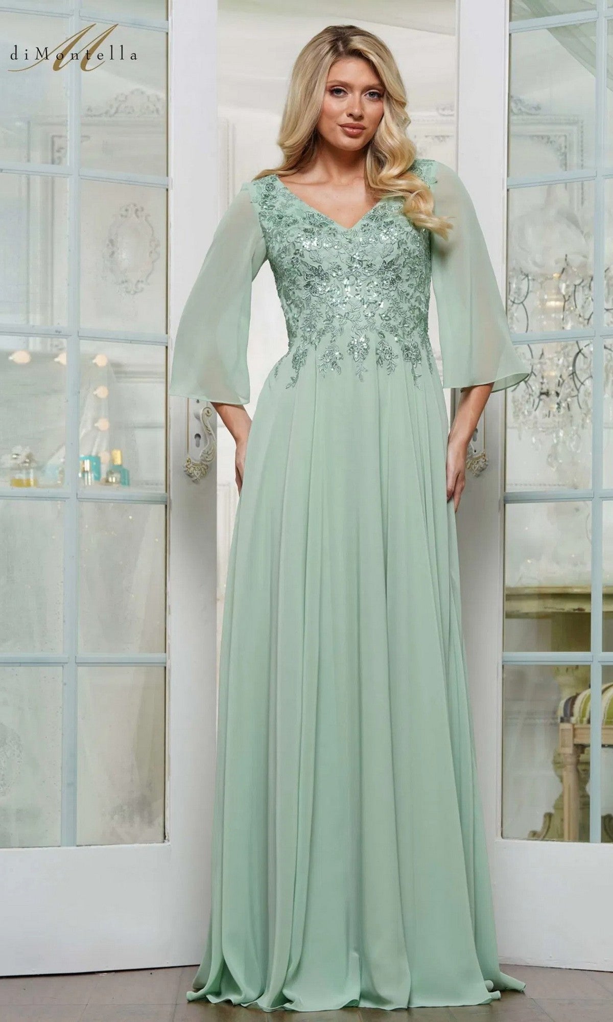 A woman in the Rina Di Montella RD3241 long formal dress in light sage, featuring sheer sleeves and a floral-embroidered bodice, stands before glass-paneled doors.